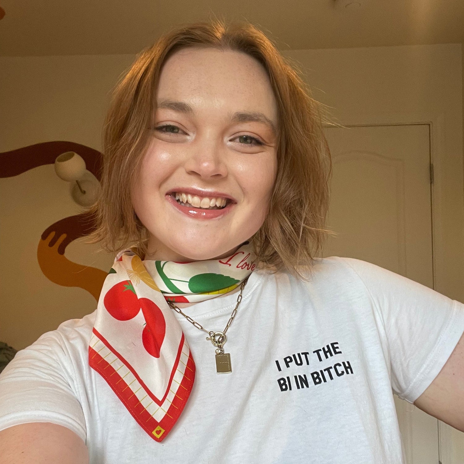 Franki smiles at the camera. She has shoulder-length blonde hair and is wearing a red and white scarf around her neck. Her t-shirt reads "I put the bi in bitch." 